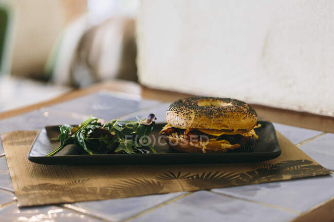 Sándwich de rosquillas con queso y pollo servido en el plato con ensalada de rúcula en la mesa en la cafetería - foto de stock