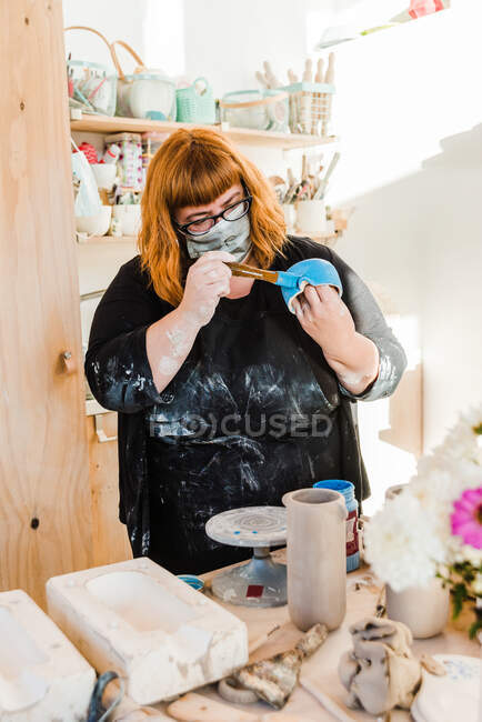 Unrecognizable woman in mask and dirty black apron standing table with pottery wheel and clay vase near forms for pottery while painting handmade ceramic pot with paintbrush in workshop — Stock Photo