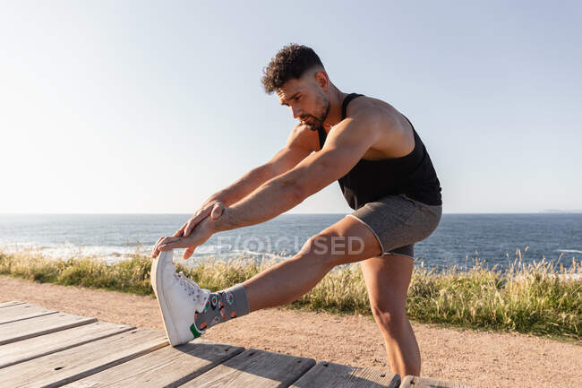Side view of muscular male doing forward bends and warming up before workout while standing near promenade on seashore — Stock Photo