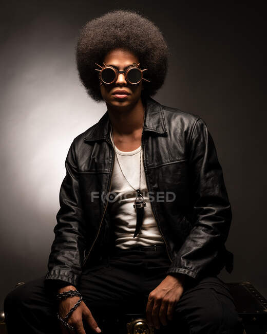 Модный афроамериканец в кожаной куртке и сапогах сидит на сером фоне — стоковое фото