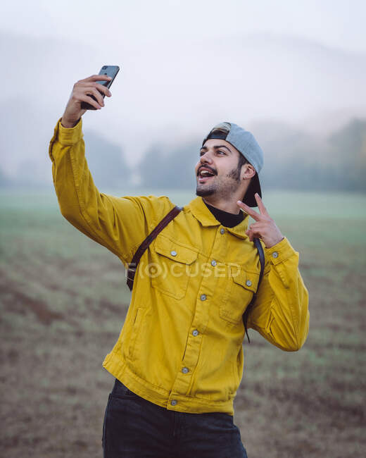 Усміхнений молодий чоловік рюкзак в жовтій джинсовій куртці бере селфі на смартфон, стоячи на туманному лузі в природі — стокове фото
