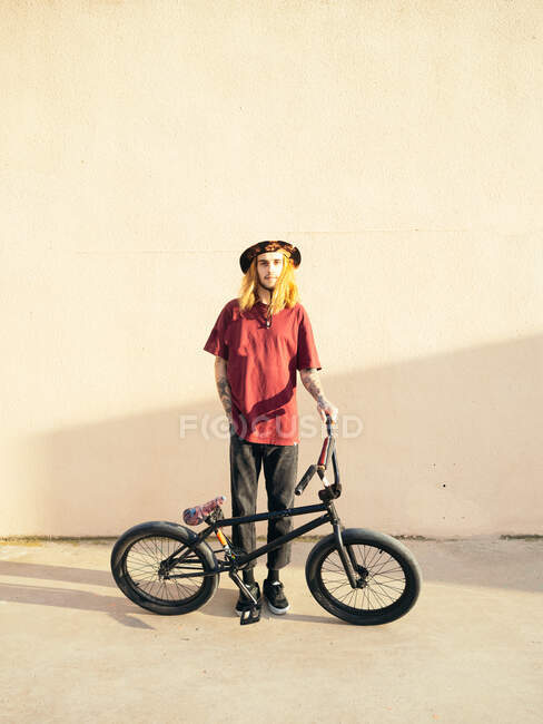 Joven atleta hipster masculino con la mano en el bolsillo y la bicicleta de prueba mirando a la cámara en la pasarela - foto de stock