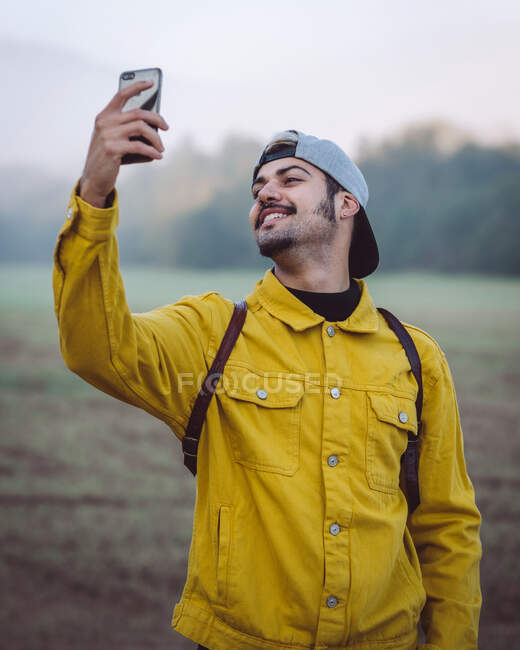 Sorrindo jovem mochileiro masculino em casaco de ganga amarelo tirando selfie no smartphone enquanto estava em pé no prado nebuloso na natureza — Fotografia de Stock