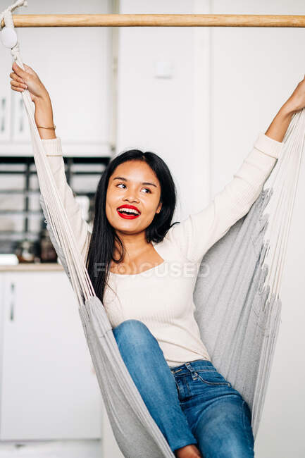 Junge positive ethnische Frau sitzt in der Hängematte und hebt die Hände, während sie zu Hause lacht und ruht — Stockfoto
