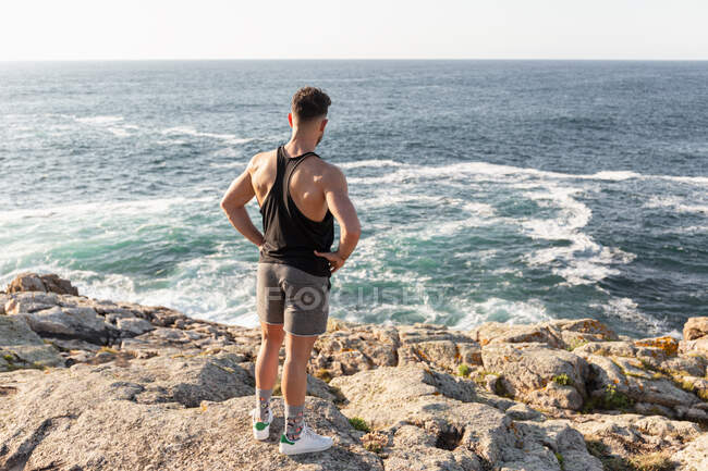 Vista posterior de atleta masculino muscular en ropa deportiva de pie en la playa y disfrutando de la puesta del sol después del entrenamiento en verano - foto de stock