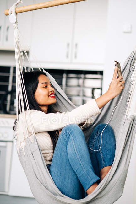 Mujer étnica feliz con sonrisa dentada sentado en hamaca con auriculares y tener videollamada en el teléfono móvil - foto de stock