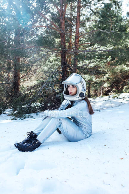 Seitenansicht der fokussierten jungen Astronautin in Raumanzug und Helm, die wegschaut und im verschneiten Wald sitzt — Stockfoto