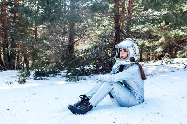 Side view of focused young female astronaut in spacesuit and helmet looking away and sitting in snowy woodland — Stock Photo