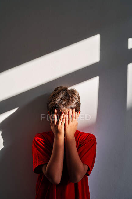 Aufgebrachter, unkenntlich gemachter Junge bedeckt Gesicht mit Händen und weint, während er in der Nähe der Mauer zu Hause steht — Stockfoto