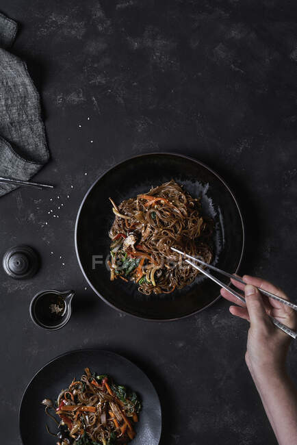 Top view of crop unrecognizable person taking food with chopsticks from plate with Korean dish Japchae cooked from noodles and vegetables — Stock Photo
