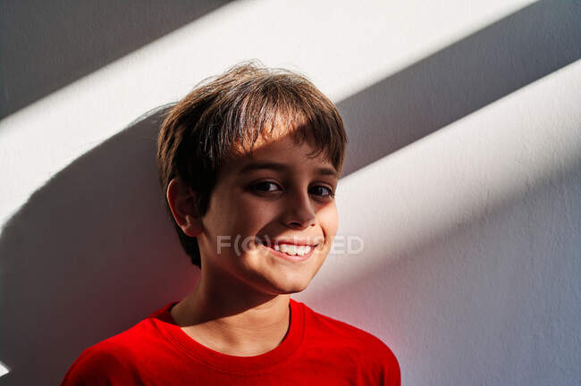 Alegre menino pré-adolescente olhando para a câmera enquanto estava em pé no fundo da parede branca — Fotografia de Stock