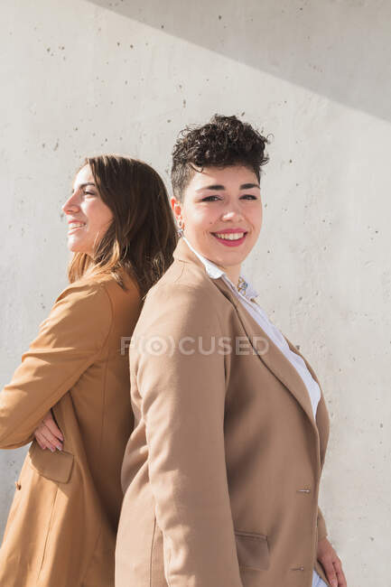 Vista laterale di giovani donne sorridenti in abiti eleganti in piedi schiena a schiena, mentre guardando lontano nella giornata di sole vicino al muro grigio — Foto stock