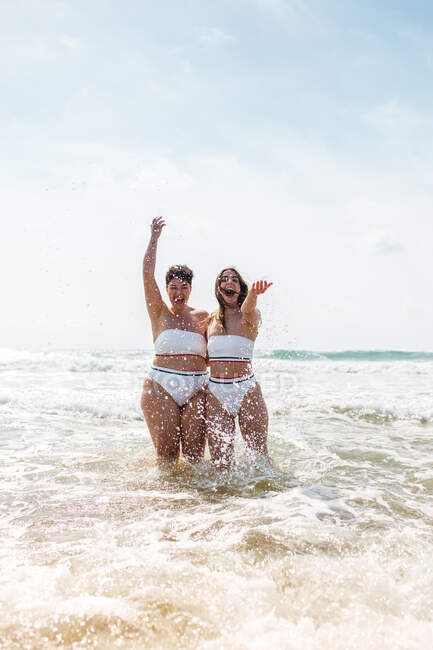 Веселі друзі-жінки в купальниках обіймаються один з одним, стоячи бризкаючи воду в піщаному океані біля піщаного пляжу під блакитним хмарним небом в сонячний день — стокове фото