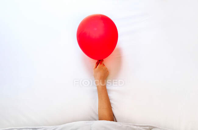 Обрезанный неузнаваемый уставший человек с красным воздушным шаром в руке спит в постели с белыми простынями после вечеринки — стоковое фото