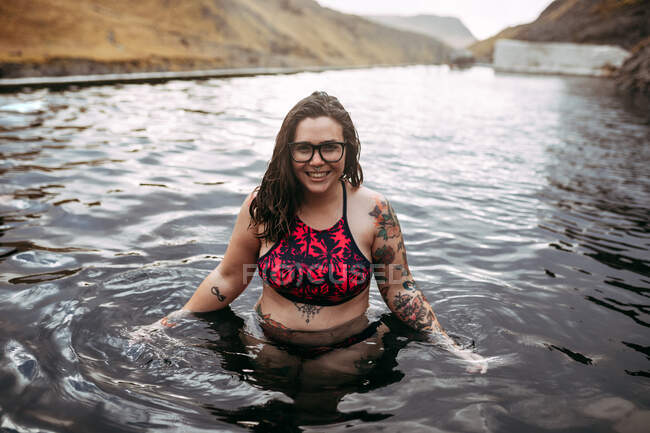 Jovem alegre tatuado hipster em maiô na água entre as montanhas — Fotografia de Stock