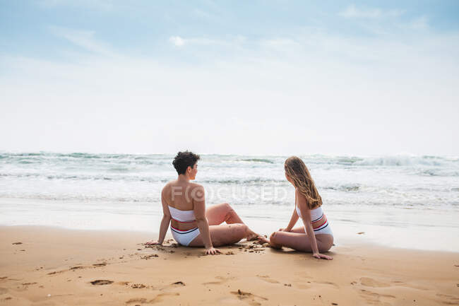 Visão traseira de jovens amigas sorridentes irreconhecíveis em trajes de banho brancos sentados na costa arenosa perto do oceano sob o céu azul nublado em dia ensolarado — Fotografia de Stock
