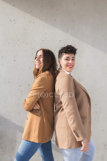 Вид сбоку улыбающихся молодых женщин в стильной одежде, стоящих спиной к спине и отводящих взгляд в солнечный день у стены — стоковое фото