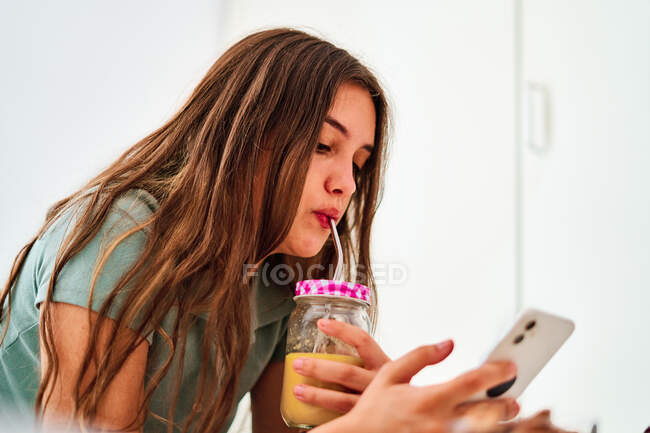 Вид збоку молодої студентки, яка переглядає соціальні мережі на мобільному телефоні біля столу зі свіжими фруктами та соком, проводячи вдома вранці — стокове фото