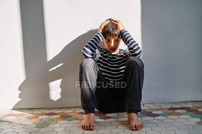 Niño infeliz sentado en el suelo y cubriendo la cabeza con las manos mientras sufre de violencia doméstica en el hogar - foto de stock