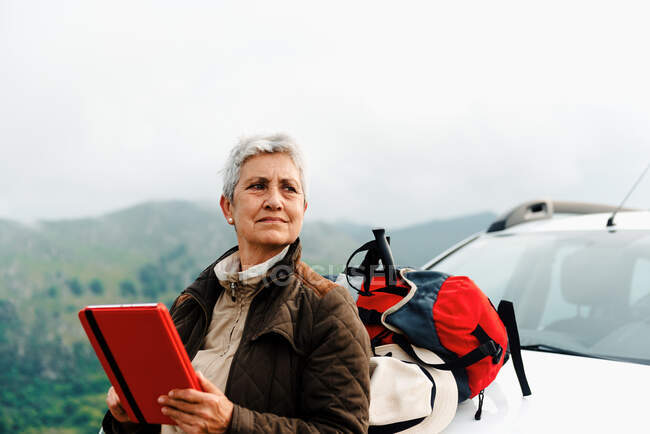 Пожилая женщина-путешественница с короткими седыми волосами, опираясь на автомобиль и просматривая планшет во время дорожной поездки в сельской местности и глядя в сторону — стоковое фото