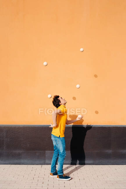 Вид сбоку на все тело молодого мужчины, выполняющего трюк с жонглирующими шарами, стоя на тротуаре возле ярко-оранжевой стены — стоковое фото