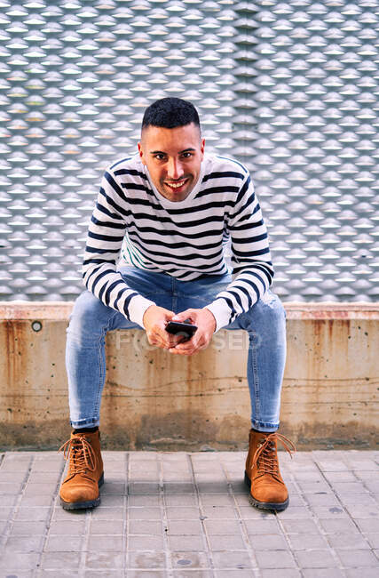 Homem hispânico feliz em roupas casuais olhando para a câmera com sorriso e segurando um smartphone enquanto sentado na fronteira contra a parede ornamental na rua da cidade — Fotografia de Stock