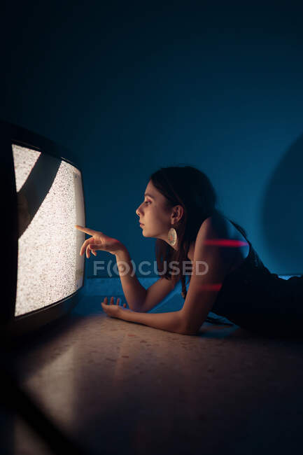Vista lateral do modelo feminino em vestido preto deitado no chão perto de uma televisão antiga brilhante no estúdio escuro — Fotografia de Stock