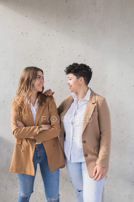 Молодые улыбающиеся женщины в стильной одежде, стоящие вместе, глядя друг на друга в солнечный день возле серой стены — стоковое фото