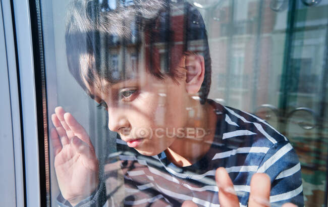 Attraverso vetro di infelice ragazzo preadolescente con lividi sul viso guardando altrove mentre in piedi vicino alla finestra a casa come concetto di violenza domestica e abusi sui bambini — Foto stock