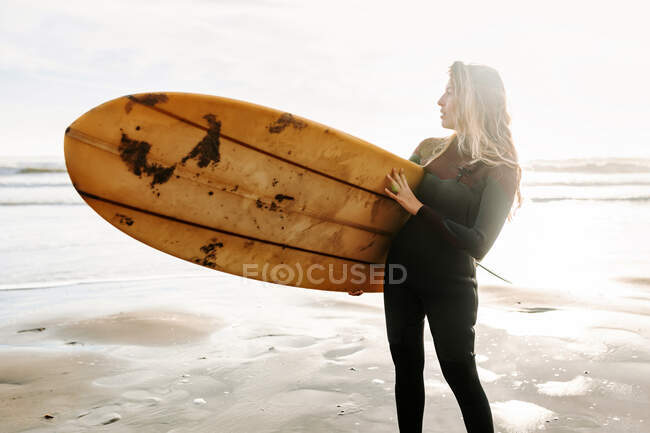 Surfer woman dressed in wetsuit standing looking away with the surfboard on the beach during sunrise in the background — Stock Photo