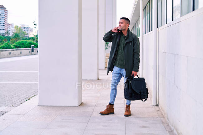 Pieno corpo ispanico uomo in abiti casual guardando altrove e parlando sul telefono cellulare mentre in piedi sul marciapiede prima del lavoro — Foto stock
