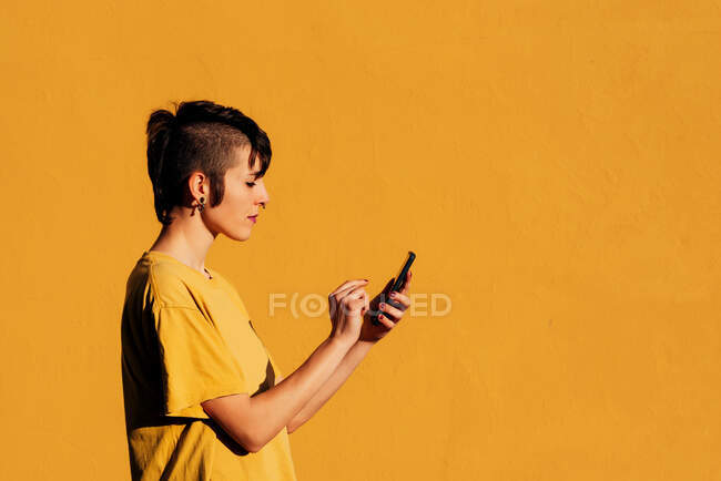 Вид збоку сучасної жінки зі стильною стрижкою та пірсингом за допомогою смартфона для текстового повідомлення в соціальних мережах на жовтому фоні — стокове фото