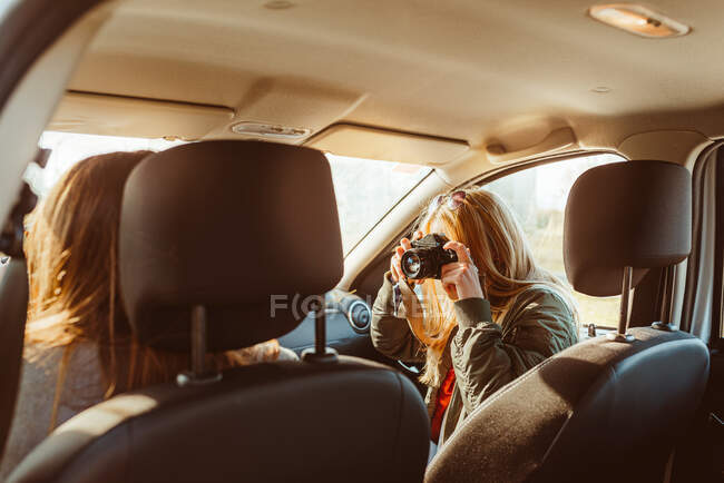 Mulher com câmera vintage fotografar amigo condução carro feminino ter viagem em dia ensolarado — Fotografia de Stock