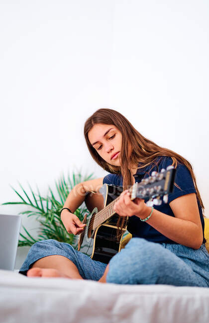 Estudante focada assistindo vídeo tutorial no laptop enquanto aprendia a tocar guitarra acústica durante o tempo livre em casa — Fotografia de Stock