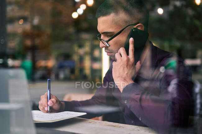 Homme d'affaires hispanique intelligent dans des lunettes prendre des notes dans le bloc-notes et répondre à l'appel téléphonique tout en étant assis derrière la fenêtre à la cafétéria — Photo de stock