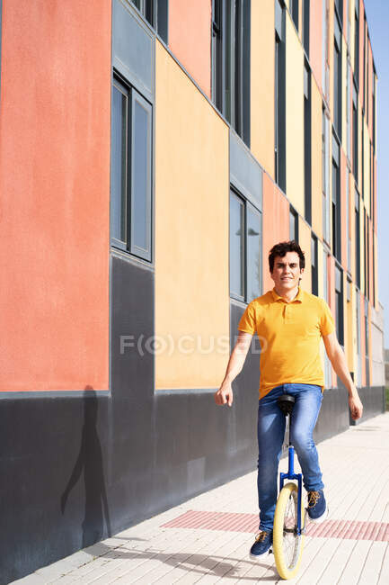 Full body of happy man in casual wear riding on unicycle on modern urban street with colorful building looking at camera — Stock Photo