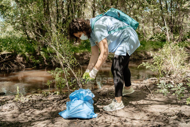 Активная женщина-эко волонтер в защитной маске и перчатках собирает мусор из ручья в мусорный мешок в летнем лесу — стоковое фото