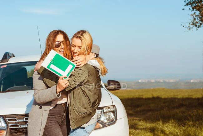 Fröhliche junge Freundinnen mit Low-Gang-Schild stehen an weißem Auto am Ufer des Meeres und umarmen sich beim Trip — Stockfoto