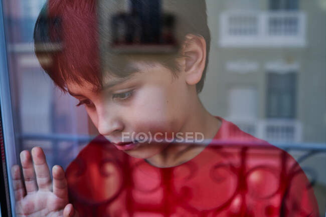 Attraverso vetro di infelice ragazzo preadolescente con lividi sul viso guardando altrove mentre in piedi vicino alla finestra a casa come concetto di violenza domestica e abusi sui bambini — Foto stock