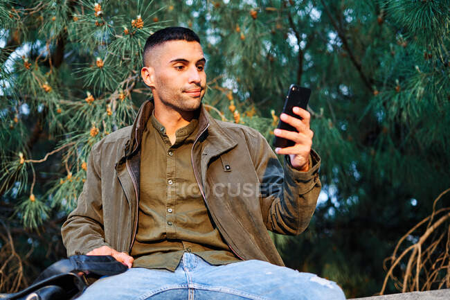 Tipo hispano en ropa casual mirando hacia otro lado y mensajes de texto en el teléfono, mientras que de pie cerca de la barrera y el árbol de coníferas en el parque - foto de stock