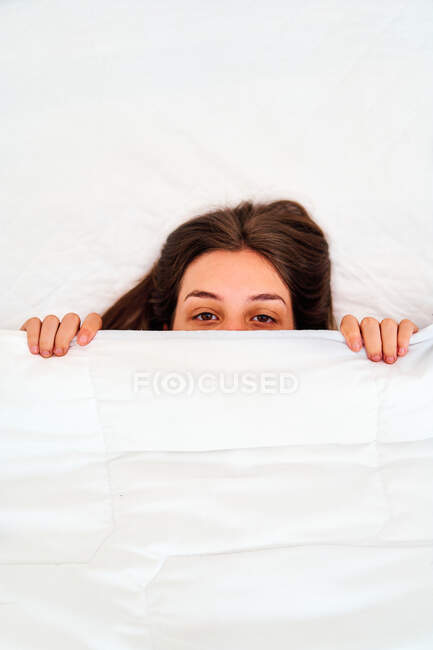 Von oben sieht eine faule junge Frau in die Kamera, die das halbe Gesicht mit weicher weißer Decke bedeckt, während sie morgens im gemütlichen Bett schläft — Stockfoto