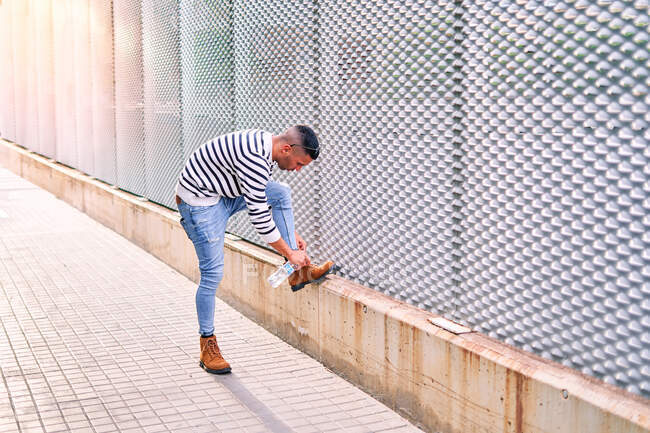 Full body guy in stylish casual outfit stepping on border near wall and tying shoelaces on boot on city street at daytime — Stock Photo