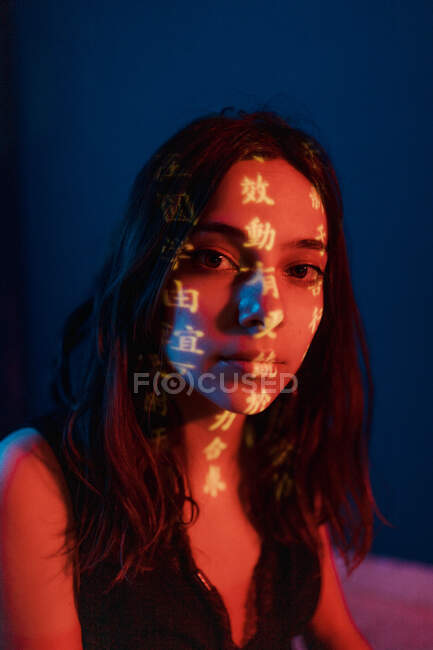 Fashionable young female model with light projection in shape of oriental hieroglyphs looking at camera in dark studio with red illumination — Stock Photo