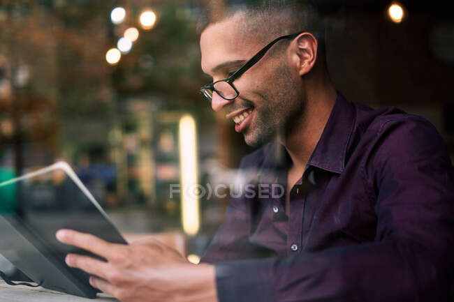 Hombre hispano inteligente en gafas leyendo notas en planificador mientras está sentado a la mesa detrás de la ventana y trabajando en un proyecto de negocios en la cafetería - foto de stock