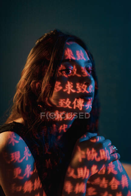 Modisches junges weibliches Modell mit Lichtprojektion in Form orientalischer Hieroglyphen, die im dunklen Atelier mit roter Beleuchtung wegschauen — Stockfoto