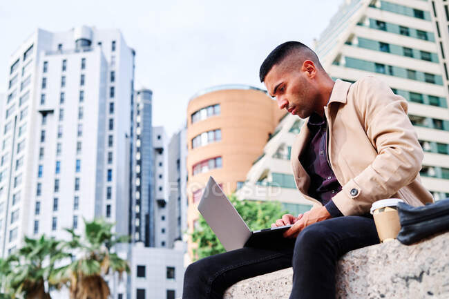 Niedriger Winkel eines fokussierten jungen hispanischen Mannes in stylischer Freizeitkleidung, der ferngesteuert am Laptop an einem freiberuflichen Projekt arbeitet, während er mit einer Tasse Kaffee in der Nähe moderner städtischer Gebäude sitzt — Stockfoto