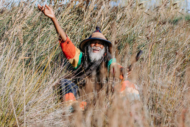 Vieux rastafari ethnique joyeux avec dreadlocks regardant la caméra debout dans une prairie sèche dans la nature — Photo de stock