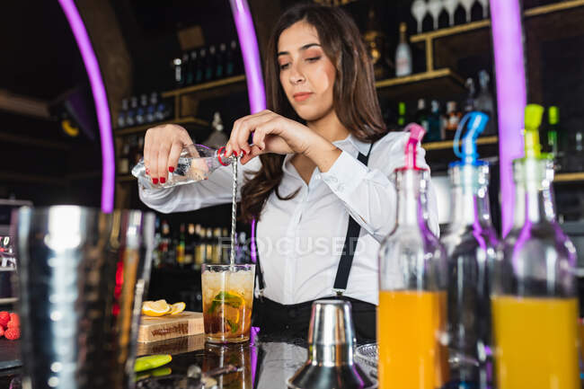 Женщина-бармен в стильном наряде добавляет жидкость из бутылки в стекло и перемешивает длинной ложкой, готовясь коктейль стоя у стойки в современном баре — стоковое фото