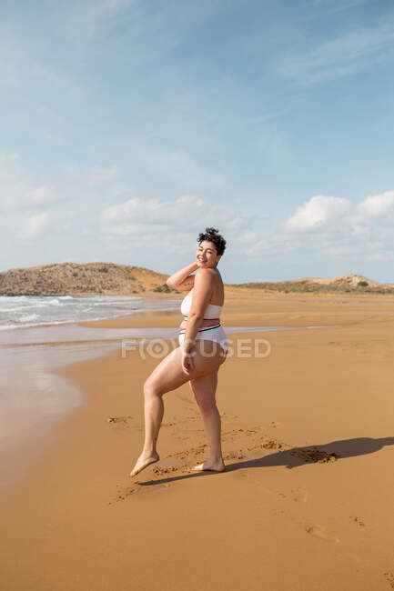 Full length of young female in swimsuit standing looking at camera on sandy coast in sunny day under blue cloudy sky — Stock Photo