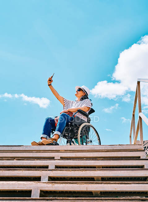 Зрелая женщина-инвалид сидит в инвалидной коляске и делает селфи на мобильном телефоне возле лестницы на фоне голубого неба в городе — стоковое фото